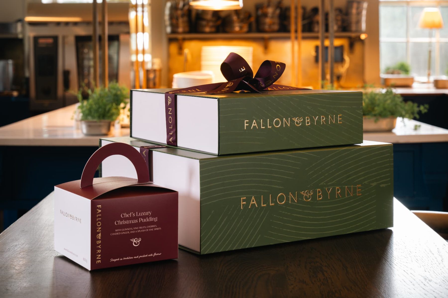 Two olive green magnetic gift boxes with gold foil Fallon and Byrne branding and burgundy printed ribbon, beside a burgundy handled pudding carton.
