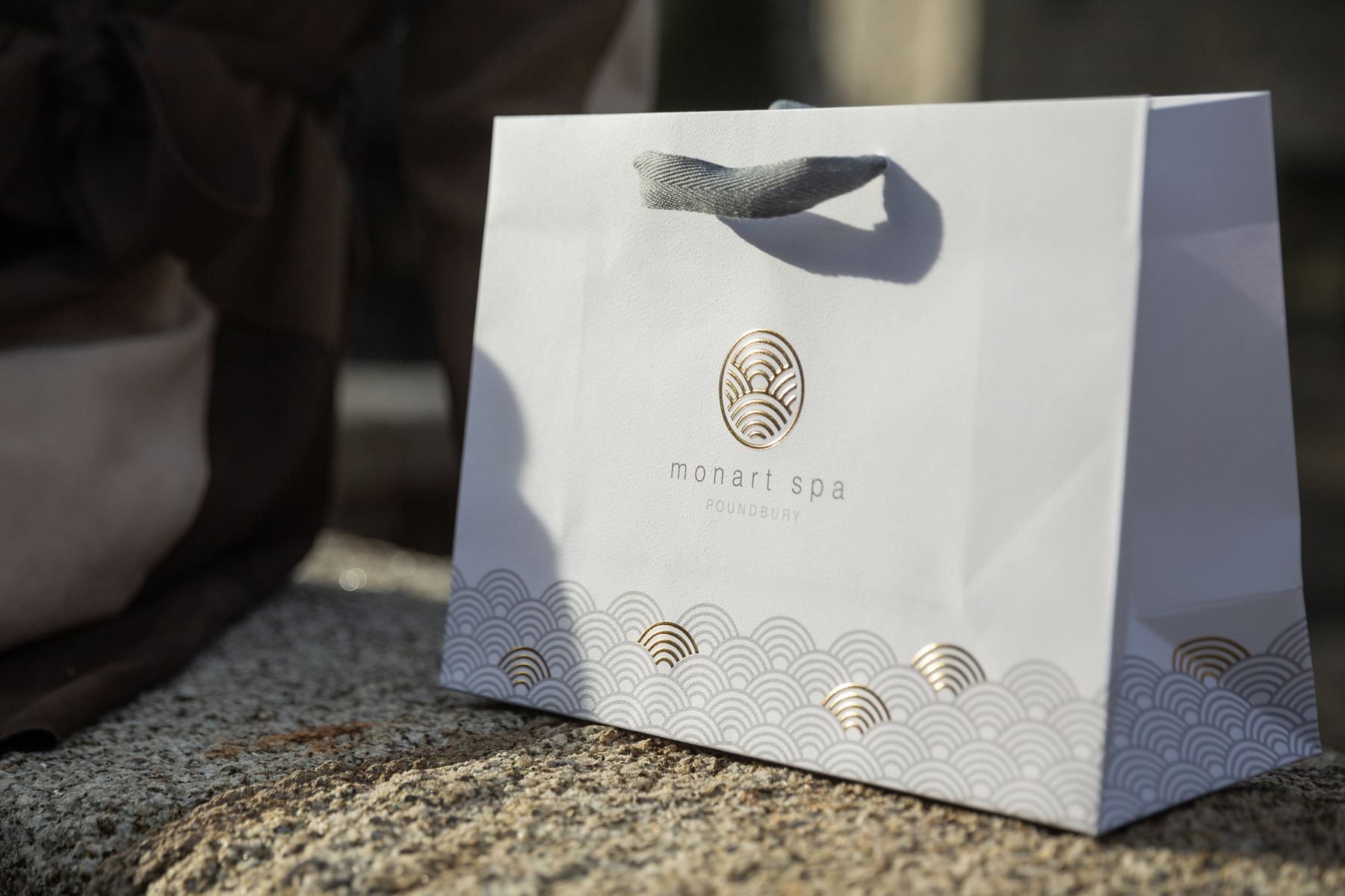 White landscape paper carrier bag with grey ribbon handles, gold foil and embossed Monart Spa logo, and a gold and grey wave pattern base.