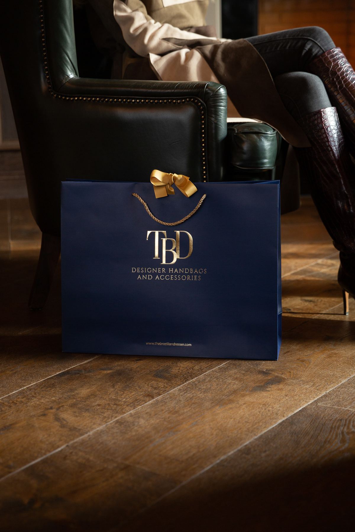 Navy blue matt laminated paper bag with gold foil 'TBD Designer Handbags and Accessories' logo, gold twisted rope handles, and a decorative gold ribbon bow.